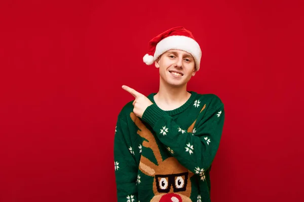 Happy guy in a Christmas sweater and hat stands on a red background and points his hands to the empty seat, looks into the camera and smiles. A smiling young man points his finger at an empty space.