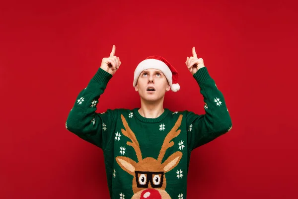 Chockad stilig ung man i julkläder och tomte hatt på röd bakgrund tittar och pekar uppåt till tomt utrymme. Förvånad kille dyker upp på kopieringsutrymme. Isolerad. — Stockfoto