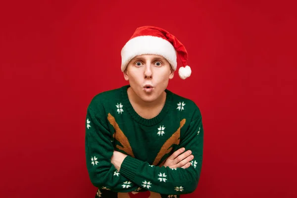 Ritratto di giovane sorpreso in cappello di Babbo Natale e caldo maglione di Natale guardando in macchina fotografica con viso scioccato su sfondo rosso. Foto di ragazzo sorpreso in abiti natalizi — Foto Stock