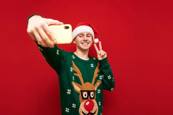 Joven feliz en ropa de Navidad tomando selfies en el teléfono inteligente, mirando el teléfono de la cámara y mostrando gesto de paz, vistiendo invierno cálido y suéter y sombrero de santa. Selfie de Navidad. Aislado . —  Fotos de Stock