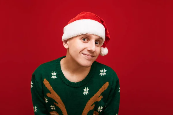 Primer plano retrato de estudio de un tipo divertido en ropa de Navidad, con un sombrero de santa y un suéter verde, mirando en cámara con una cara divertida. Chico divertido con el estado de ánimo de Navidad aislado sobre fondo rojo . —  Fotos de Stock