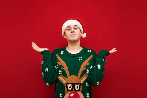 Joven divertido en sombrero de santa y suéter caliente aislado sobre fondo rojo, perplejo mirando a la cámara y extendiendo sus manos a los lados. Confundido chico en ropa de Navidad sobre fondo rojo . —  Fotos de Stock