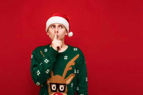 Joven divertido en sombrero de santa y suéter de Navidad se encuentra sobre un fondo rojo, muestra un signo de silencio y mira hacia otro lado en un espacio en blanco con una cara sorprendida. Aislado . —  Fotos de Stock