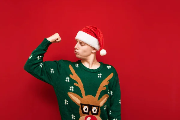 Αστείος νεαρός με χριστουγεννιάτικα ρούχα που δείχνει δικέφαλους στο κόκκινο φόντο και κοιτάζει πλάγια, φορώντας καπέλο Σάντα και ζεστό πουλόβερ. Απομονωμένοι. Αντιγραφή χώρου — Φωτογραφία Αρχείου