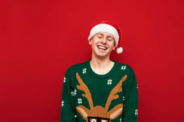 Cheerful guy stands on a red background and sincerely smiles with his eyes closed, wears a santa hat and a Christmas mood. Young man with Christmas mood isolated on red background.