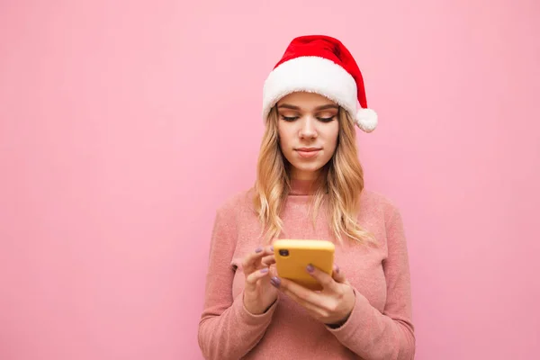 Attractive girl in pink clothes and Christmas hat isolated on a pink background, uses a smartphone with a serious face. Lady in santa hat uses internet on smartphone, isolated on pink — Stock Photo, Image