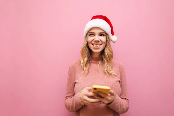 Happy girl in warm pink sweater and Santa hat stands on pink background with smartphone in hand, looks away at blank space and smiles. Christmas concept. Xmas — Stock Photo, Image
