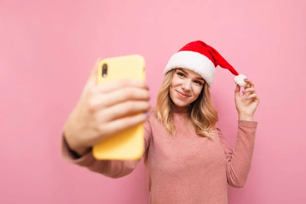 サンタの帽子の魅力的な女の子はピンクの背景に自撮りを行い、スマートフォンやポーズのカメラを調べます。クリスマスの帽子の正の女の子は、スマートフォンのカメラで休日の写真を作ります。隔離。. — ストック写真