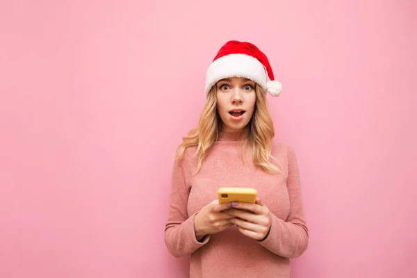 Shocked girl in christmas hat holds smartphone and looks surprised at camera on pink background. Surprised beautiful girl in pink sweater, uses smartphone, isolated. Xmas concept — Stock Photo, Image