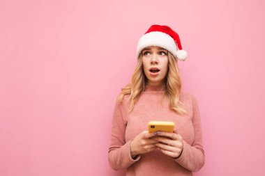 Şok olmuş pembe kazaklı ve Noel Baba şapkalı kızın portresi pembe arka planda akıllı telefon kullanıyor ve boş alana şaşırmış görünüyor, Noel şapkası takıyor. Kopyalama. X- mas konsepti