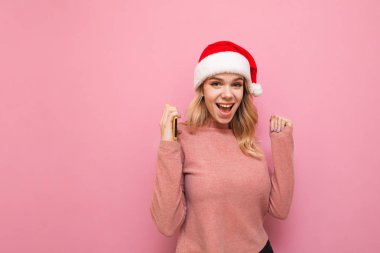 Noel Baba şapkalı mutlu bayan pembe bir arka planda duruyor ve elleri havada seviniyor, elinde bir akıllı telefon tutuyor, kameraya bakıyor ve gülümsüyor. Çekici sarışın mutlu kız Noel 'de
