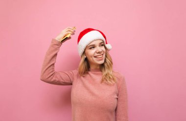 Pembe kazaklı ve Noel şapkalı gülümseyen bayan elini kaldırarak seviniyor, başka tarafa bakıyor ve pembe arka planda gülümsüyor. Mutlu sarışın bayanın portresi. Noel 'de neşeli, izole. Kopyalama alanı