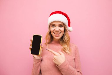 Pembe arka planda Noel Baba şapkalı mutlu bir sarışın. Elinde akıllı telefon, ekranda parmağını gösteriyor. Kameraya bakıp gülümsüyor. Akıllı telefonu gösteren Noel şapkalı hoş bir kadının portresi..
