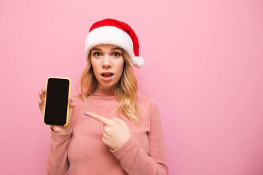 Şok olmuş, Noel şapkalı bir kız pembe arka planda duruyor, elinde bir akıllı telefon tutuyor, ekranda parmağını gösteriyor. Şaşkın kadın akıllı telefonu işaret ediyor ve kameraya bakıyor.