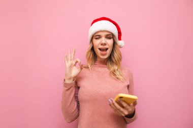 Noel şapkalı mutlu kızın portresi elinde akıllı telefonuyla pembe arka planda oturur, kameraya bakar, gülümser ve OK jestini gösterir. Akıllı telefonlu Noel Baba şapkalı oyuncu kız. X-mas