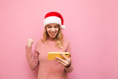 Noel şapkalı mutlu bir kızın portresi akıllı telefondan mobil oyunlar oynuyor, pembe arka planda kazanmanın sevinci, akıllı telefon ekranına bakıyor ve gülümsüyor. Noel ve Yeni Yıl kavramı.