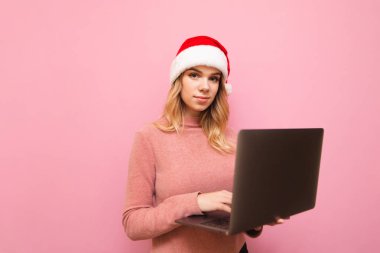 Pembe kazaklı ve Noel şapkalı bir kız pembe arka planda dizüstü bilgisayarla kameraya bakıyor ve ciddi bir yüzle kameraya bakıyor.