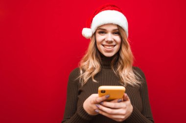 Kırmızı arka planda izole edilmiş, elinde akıllı telefonuyla kameraya bakıp gülümseyen mutlu bayan. Kırmızı arka planda akıllı telefonu olan gülümseyen Noel kızının yakın plan portresi..