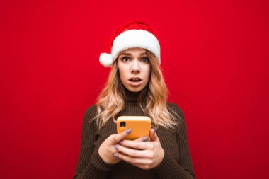 Noel şapkalı şaşkın kız elinde akıllı telefonuyla kırmızı arka planda duruyor. Şok olmuş bir şekilde kameraya bakıyor. Şok olmuş Noel Baba şapkalı kadın elinde akıllı telefonuyla kırmızı arka planda izole edilmiş..