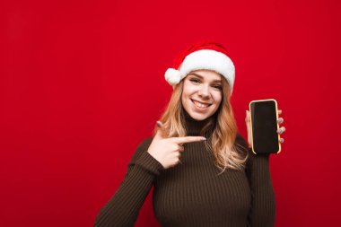 Mutlu bir kız elinde akıllı telefonuyla kırmızı bir arka planda durur ve siyah ekranda parmağını gösterir, kameraya bakar ve gülümser, bir Noel şapkası ve kalın bir kazak giyer.