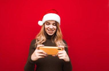 Kırmızı arka planda elinde akıllı telefonla Noel şapkası ve süveteri olan mutlu kız komik videolar izleyip gülümsüyor. Kırmızı arka planda izole edilmiş gülümseyen oyuncu kız portresi.