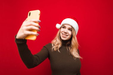 Kızaklı Noel Baba şapkalı, gülümseyen çekici kız kırmızı arka planda özçekim yapıyor, akıllı telefona bakıyor ve gülümsüyor. Sıcak süveter ve Noel şapkalı çekici bayan akıllı telefon kamerası için poz veriyor. İzole edilmiş