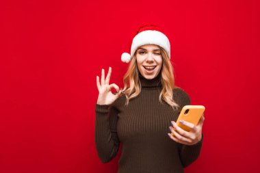 Normal giyinmiş mutlu kız elinde bir akıllı telefonla kırmızı arka planda duruyor, kameraya bakıyor ve gözünü kırpıyor, bir Noel Baba şapkası ve kalın bir kazak giyiyor. İzole edilmiş.
