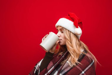 Noel şapkalı çekici sarışın kız kırmızı arka plandaki büyük bardaktan sıcak içecekle gözleri kapalı şarkı söylüyor, sıcak ekose giyiyor. Kış yılbaşı hademesi kız sıcak içki içer. Xmas