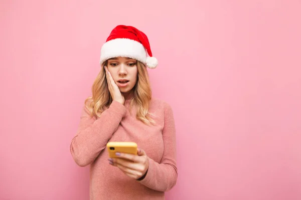 Portrait of surprised girl in pink sweater and Santa hat uses smartphone on pink background, looks into screen with surprised face. Emotional girl uses smartphone at Christmas. Isolated. Xmas concept — Stock Photo, Image