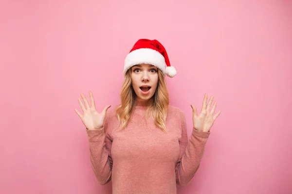 Geschokt aantrekkelijk meisje in santa hoed stak haar handen op in verbazing, kijkend in de camera met bang gezicht. Portret van een geschokte dame met kerstmuts. Kerstverkoop. Omg. — Stockfoto