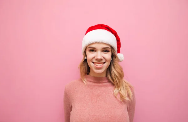 Retrato de una chica feliz con un sombrero de Santa Claus parado sobre un fondo rosa, escucha música en auriculares inalámbricos y mira a la cámara con una sonrisa en su rostro. Chica escuchando música navideña —  Fotos de Stock