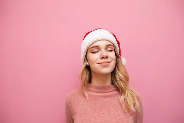 Tevreden aantrekkelijke blonde meisje in santa hoed luisteren naar muziek in gesloten-lus draadloze hoofdtelefoon en glimlachende, close-up portret. De dame met de kerstmuts luistert graag naar muziek. Kopieerruimte — Stockfoto