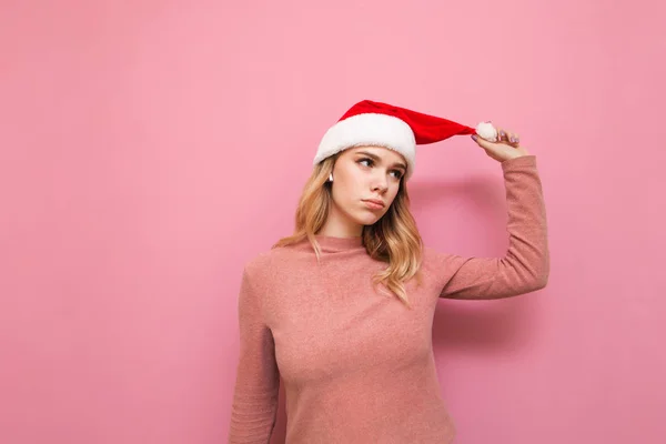 Attraktiv tjej i tomte hatt och rosa tröja isolerad på rosa bakgrund, ser bort med allvarliga ansikte. Jul inte glad flicka står på en rosa bakgrund. — Stockfoto