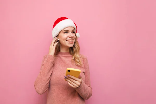 Attractive happy girl in Christmas and hat stands with smartphone in hand, listens to music in wireless headphones, smiles and looks away at copyspace. Isolated on pink background. Christmas concept. — Stock Photo, Image