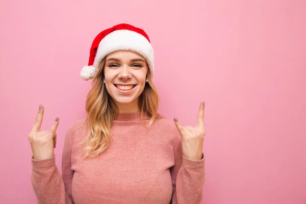 Retrato de señora feliz en sombrero de navidad escuchando música en auriculares y mostrando las manos gesto Rock y fondo rosa, mirando a la cámara y sonriendo. Aislado . — Foto de Stock