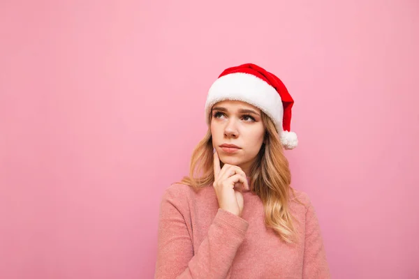 Närbild porträtt av en söt flicka i en jul hatt isolerad på en rosa bakgrund, eftertänksamt tittar bort med ett allvarligt ansikte. Pensiv dam i tomte hatt tittar på tomt utrymme. — Stockfoto