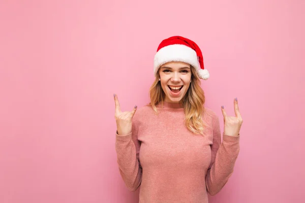 Rubia feliz en un suéter rosa y sombrero de Navidad se coloca sobre un fondo rosa, escucha música en auriculares inalámbricos y muestra una lata de metal pesado. Señora positiva en el sombrero de Navidad muestra gesto de roca . —  Fotos de Stock