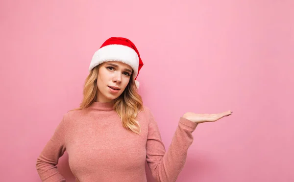 Förvirrad flicka i en julhatt står på en rosa bakgrund och håller en kopia utrymme på handen, ser in i kameran med ett överraskat ansikte. Töm sätet på en attraktiv flickas arm. — Stockfoto