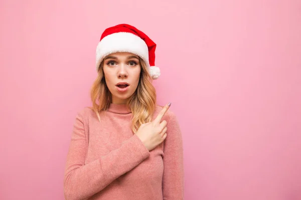 Porträtt av överraskad flicka i jul hatt och rosa tröja står på rosa bakgrund, pekar fingret på det tomma utrymmet och ser med chockad ansikte in i kameran. Julförsäljning. — Stockfoto