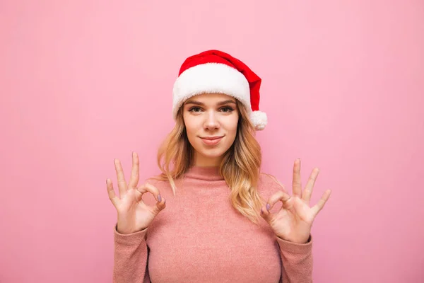 Мила дівчина в рожевому светрі і різдвяний капелюх стоїть на рожевому фоні, дивиться на камеру і показує її руки ОК жест. Гарна дівчинка в капелюсі Санта Клауса показує жест добре, ізольовано. — стокове фото