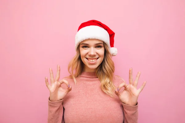 Signora sorridente in cappello di Babbo Natale e maglione rosa isolato su sfondo rosa, guarda nella fotocamera, mostra pollici in su OK e ride. Concetto natalizio. Natale. — Foto Stock