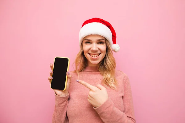 手にスマートフォンを持ってピンクの背景にサンタクロースの帽子の幸せなブロンドは、暗い画面で指を示し、カメラと笑顔を見ています。スマートフォンを示すクリスマスの帽子のかわいい女性の肖像. — ストック写真