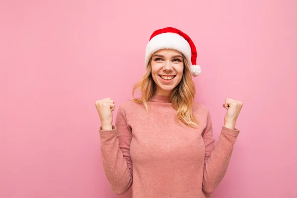 Πορτρέτο του χαρούμενου κοριτσιού σε χριστουγεννιάτικο καπέλο απομονώνονται σε ροζ φόντο, κοιτάζοντας μακριά και χαμογελώντας. Ευτυχισμένο κορίτσι χαίρεται τη νίκη με σηκωμένα χέρια. Χριστουγεννιάτικη ιδέα. Απομονωμένοι. Χριστούγεννα — Φωτογραφία Αρχείου