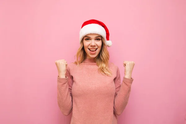 Ragazza felice gioisce nella vittoria con le braccia alzate su sfondo rosa, guarda in macchina fotografica e indossa il cappello di Natale. La signora col cappello di Babbo Natale è gioiosa. Isolato. Natale. — Foto Stock