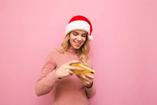 Positive blonde girl in Christmas hat plays mobile games on smartphone, looks at screen and smiles, isolated on pink background. Happy lady teenager in santa hat playing on smartphone. — Stock Photo, Image