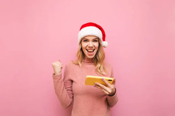 Chica rubia alegre en sombrero de santa se regocija en la victoria en el juego móvil contra el fondo rosa, se para con el teléfono inteligente en la mano, levantó la mano de alegría, mira a la cámara y sonríe. Juegos de Navidad . —  Fotos de Stock