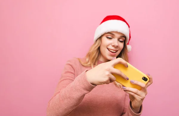 Focus sur le smartphone dans les mains d'une gamer girl.Positive adolescente blonde chapeau de Noël joue à des jeux mobiles sur smartphone, regarde l'écran et sourit, isolé sur fond rose — Photo