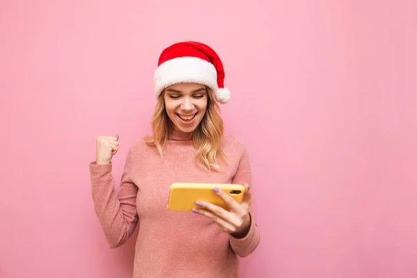 Portrait of happy girl in christmas hat playing mobile games on smartphone, joyful rejoices to win on pink background, looks at smartphone screen and smiles. Christmas and New Year concept. — Stock Photo, Image