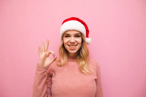 Happy Christmas girl shows OK gesture, looks in camera and rejoices on pink background. Teen girl in santa claus hat shows OK sign and poses at camera. Copy space