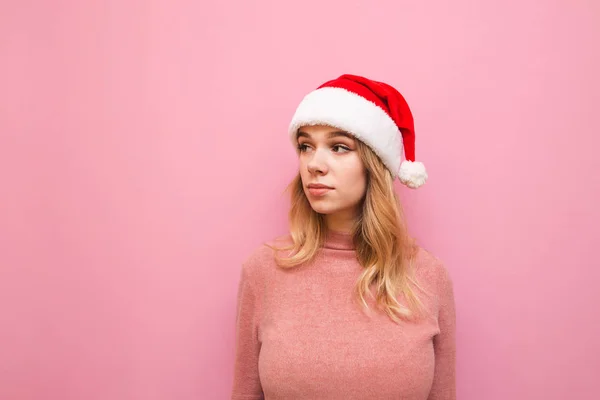 Mooi meisje in kerstmuts en roze trui kijkt weg met serieus gezicht geïsoleerd op roze achtergrond. Kerst portret van een schattig meisje in een kerstmuts. X-mas — Stockfoto